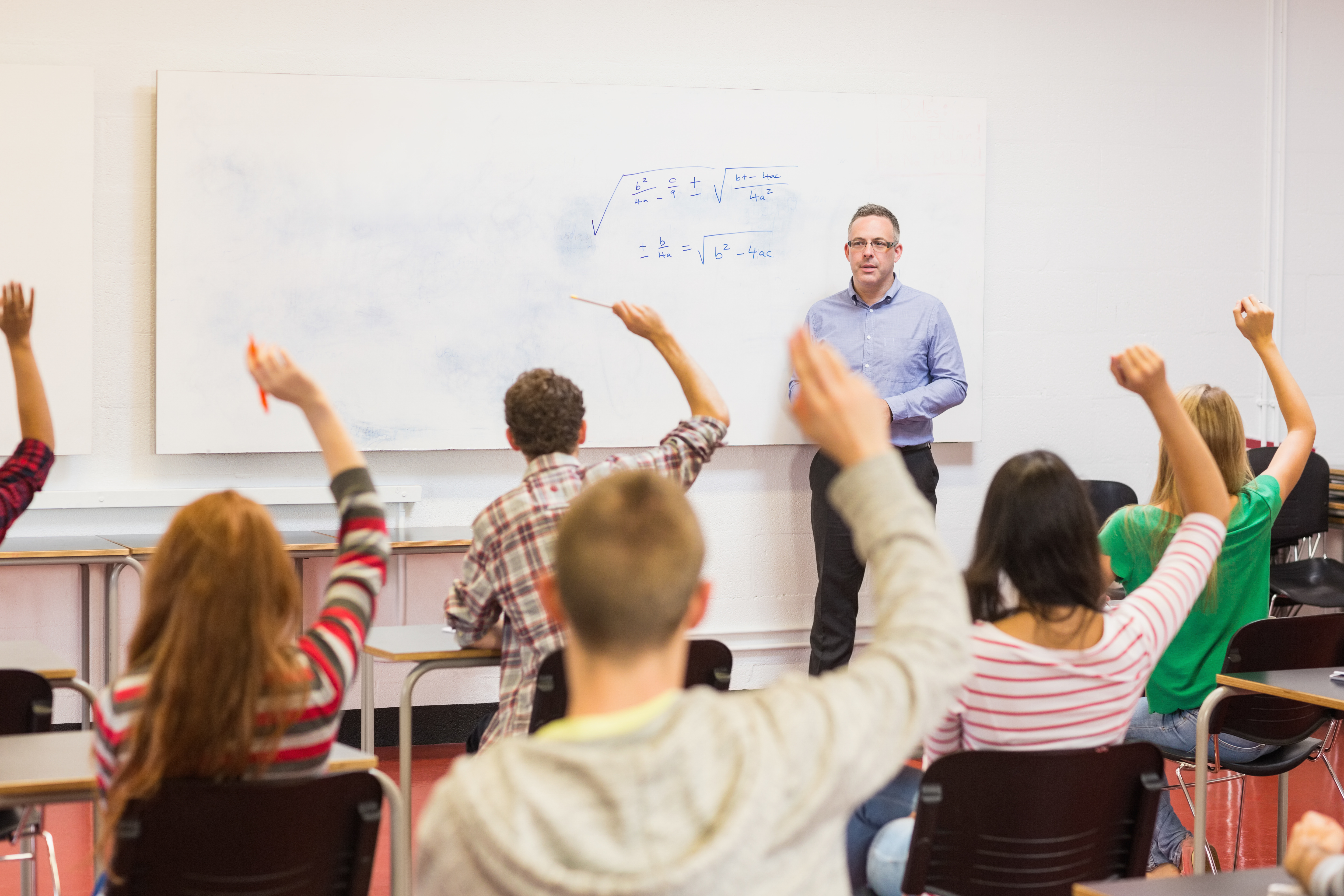 Rear view of students with hands raised with a teacher in the classroom Rear view of students with hands raised with a teacher in the classroom